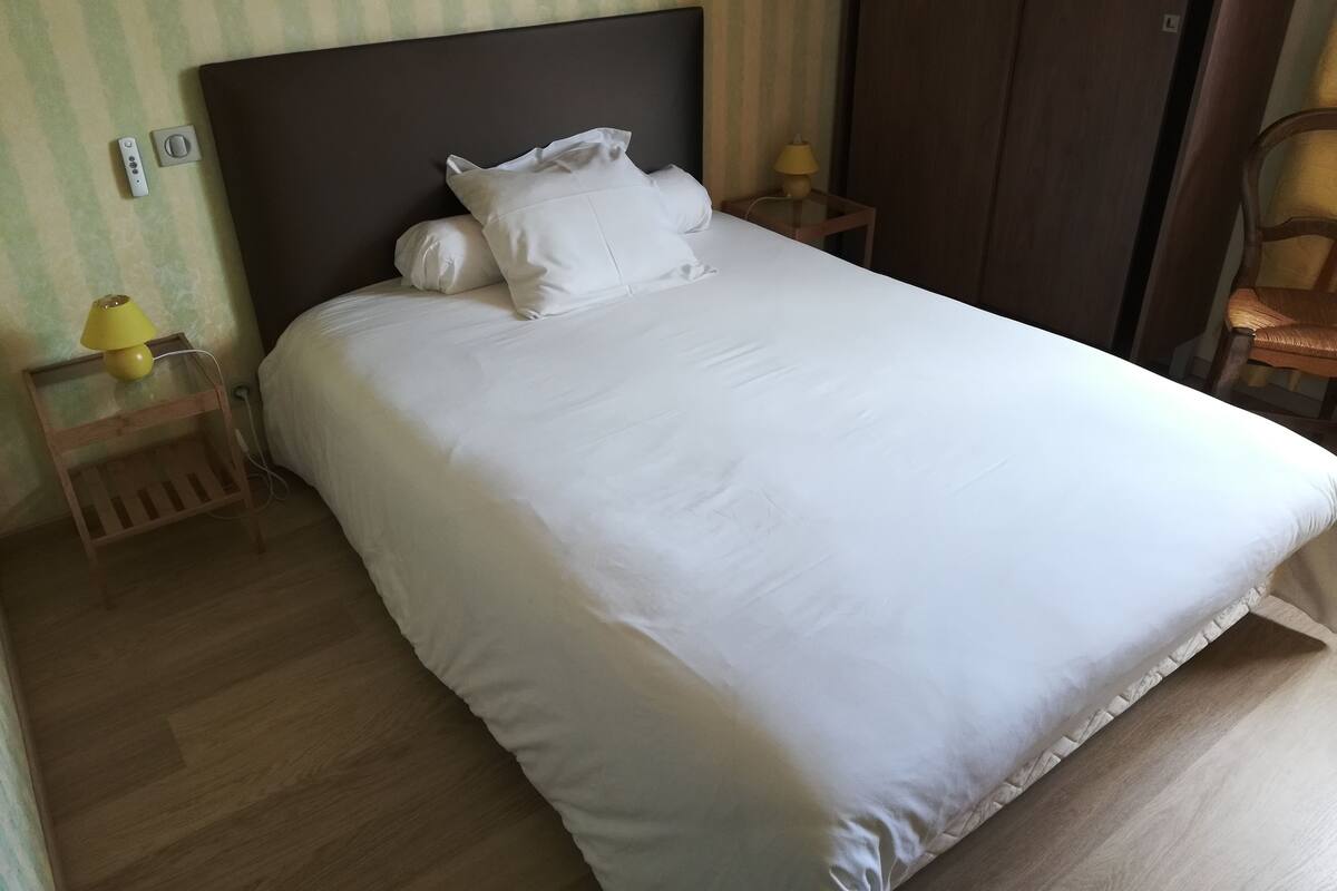 A neatly made bed with white bedding, a wooden nightstand with a lamp, and a chair.