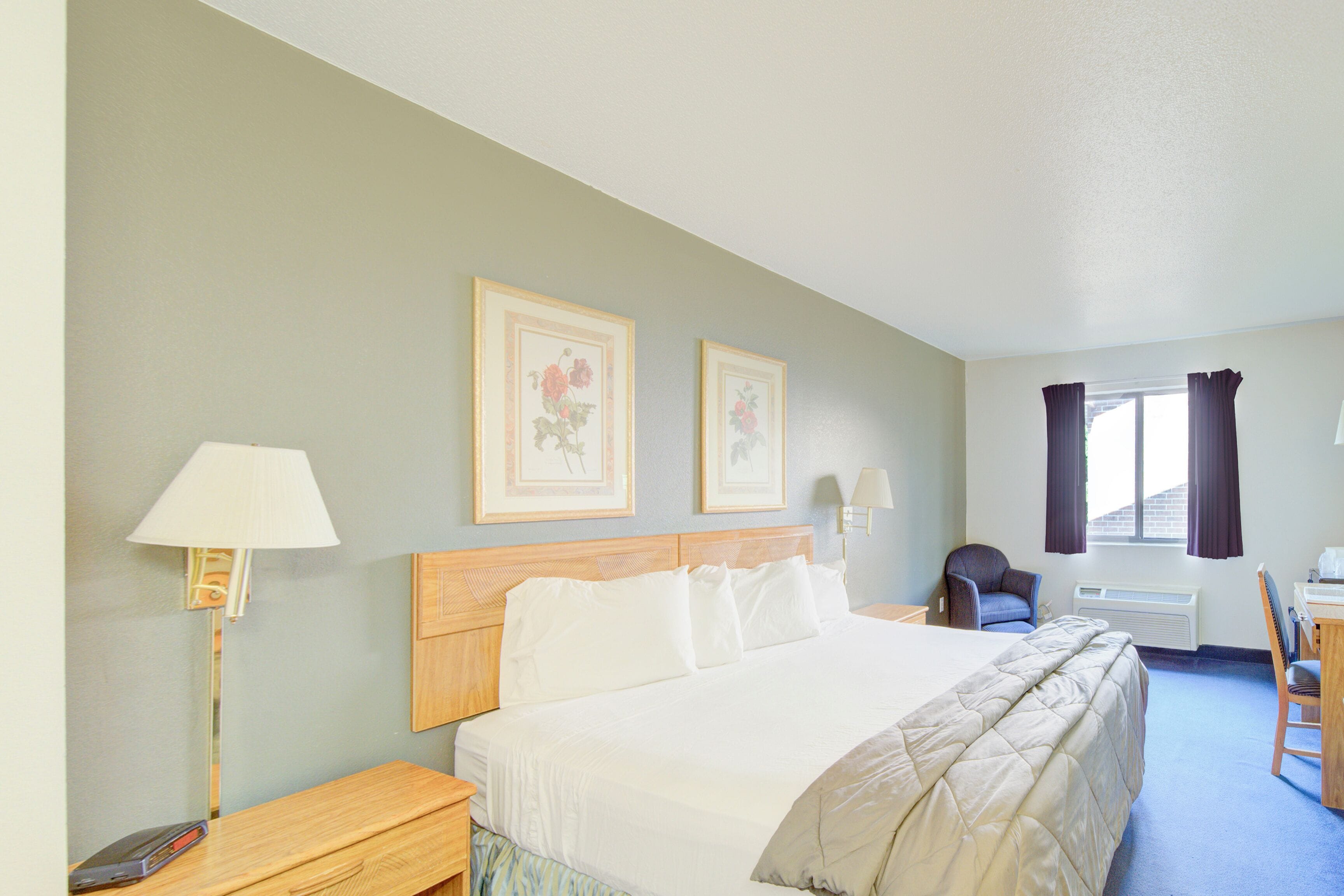 A hotel room with a large bed, a desk, a chair, a lamp, and two framed pictures on the wall.