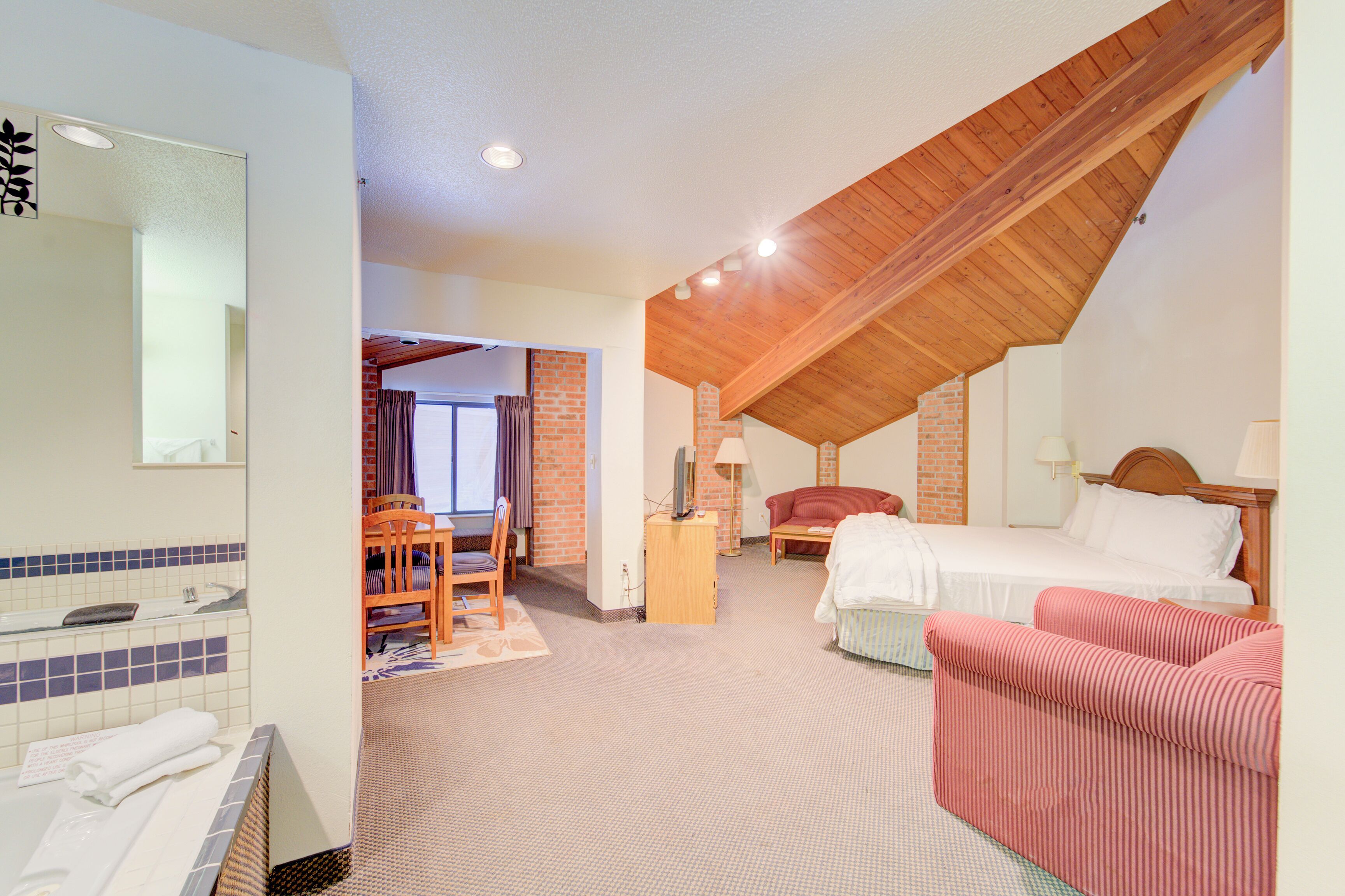 A hotel room with a bed, a chair, a desk, a mirror, and a bathroom with a bathtub and sink.