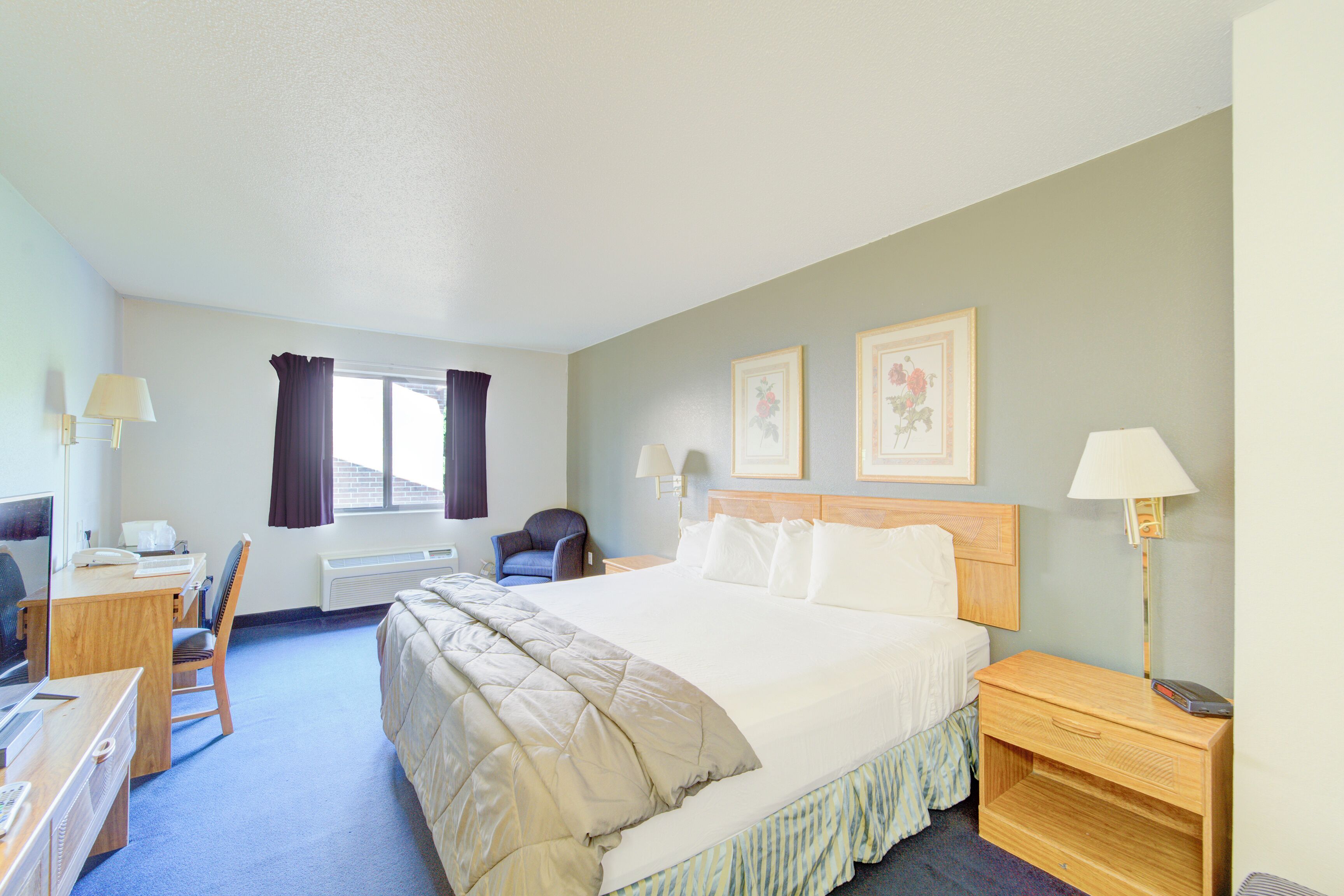 A hotel room with a large bed, a desk with a chair, a lamp, and two framed pictures on the wall.