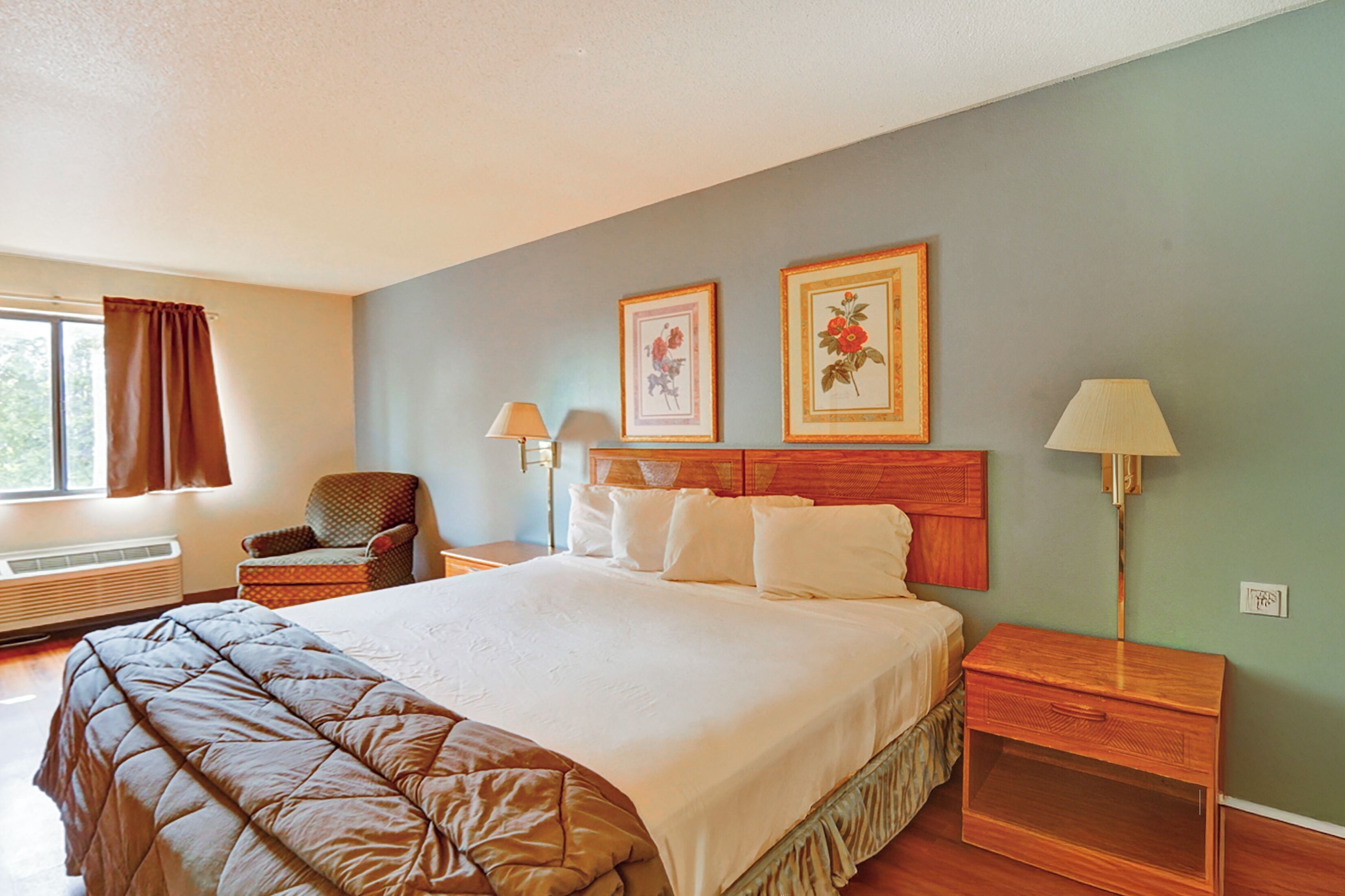 Une chambre d’hôtel bien rangée, avec un grand lit, une chaise, deux lampes de chevet et deux tableaux représentant des motifs floraux, encadrés et accrochés au mur.