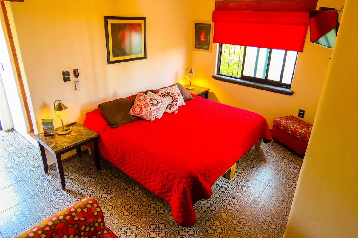 A room with a large bed, a bedside table with a lamp, a small table with a lamp, a window with a red blind, and patterned floor tiles.