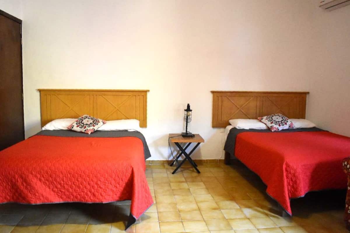 A room with two beds, each with a wooden headboard and red bedding. There is a small table with a lamp between the beds.