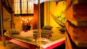 Una hamaca, una cama con almohadones estampados, una planta en maceta y una ventana iluminada con velas.