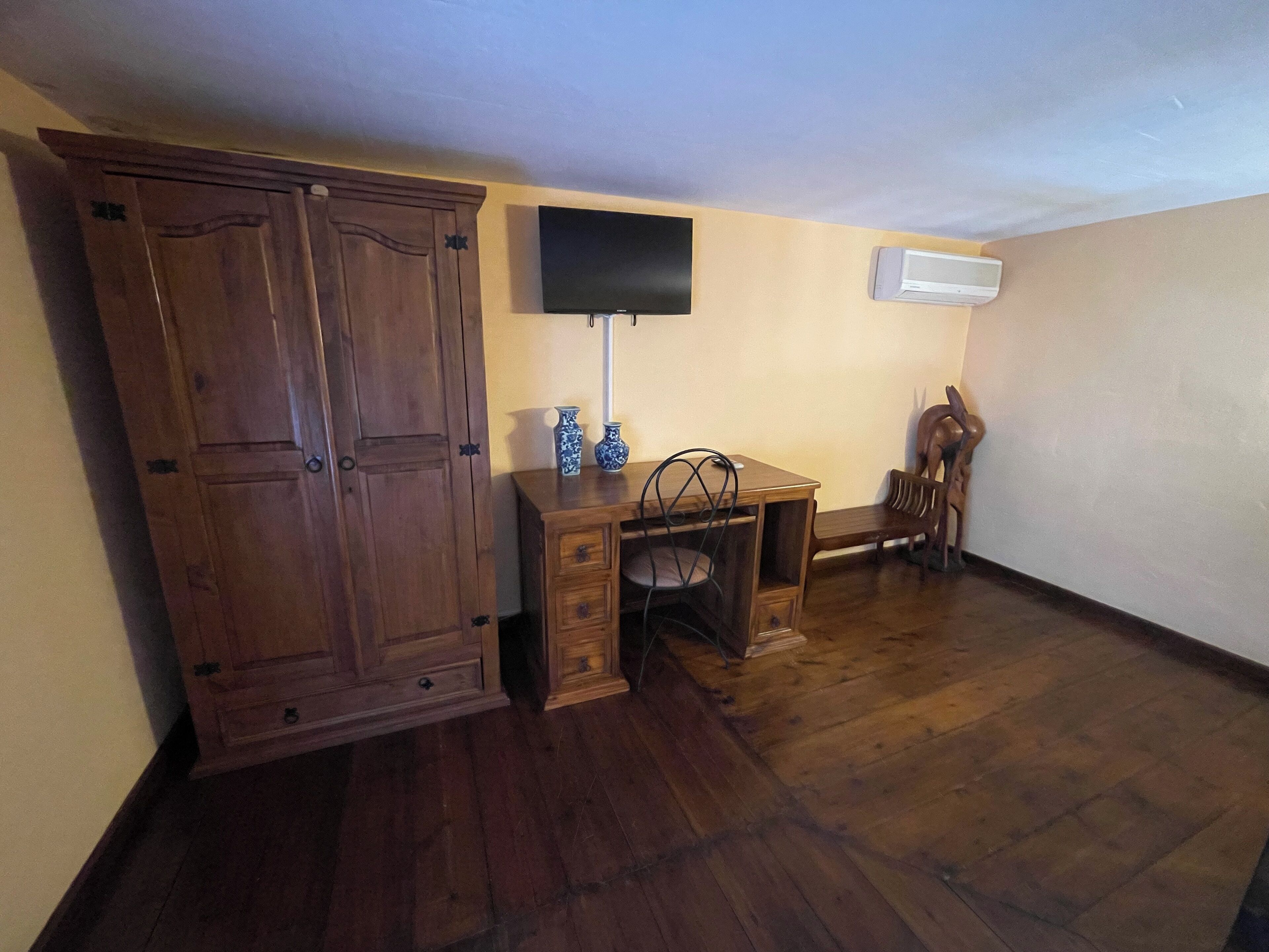A room with a wooden wardrobe, a desk with a chair, a flat-screen TV mounted on the wall, and an air conditioning unit.