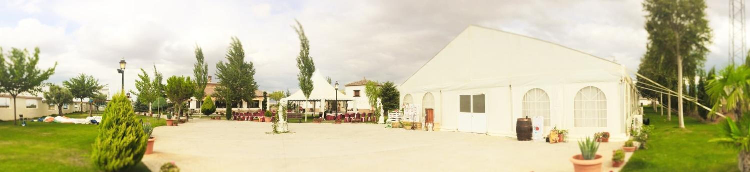 Un edificio grande y blanco con ventanas arqueadas y techo plano, rodeado de vegetación y otras construcciones.