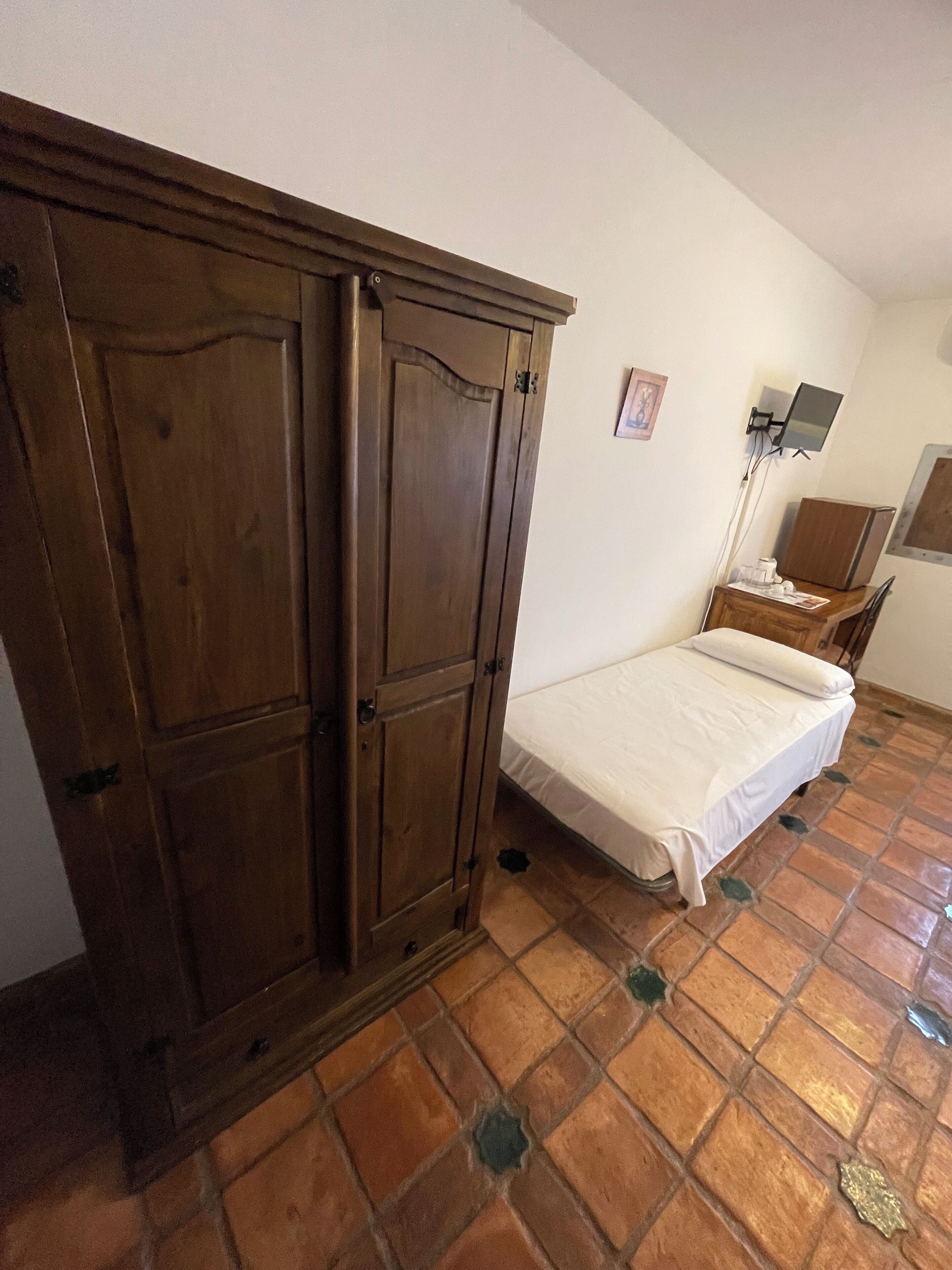 Una habitación con cama, armario, escritorio con silla y televisor de pared.