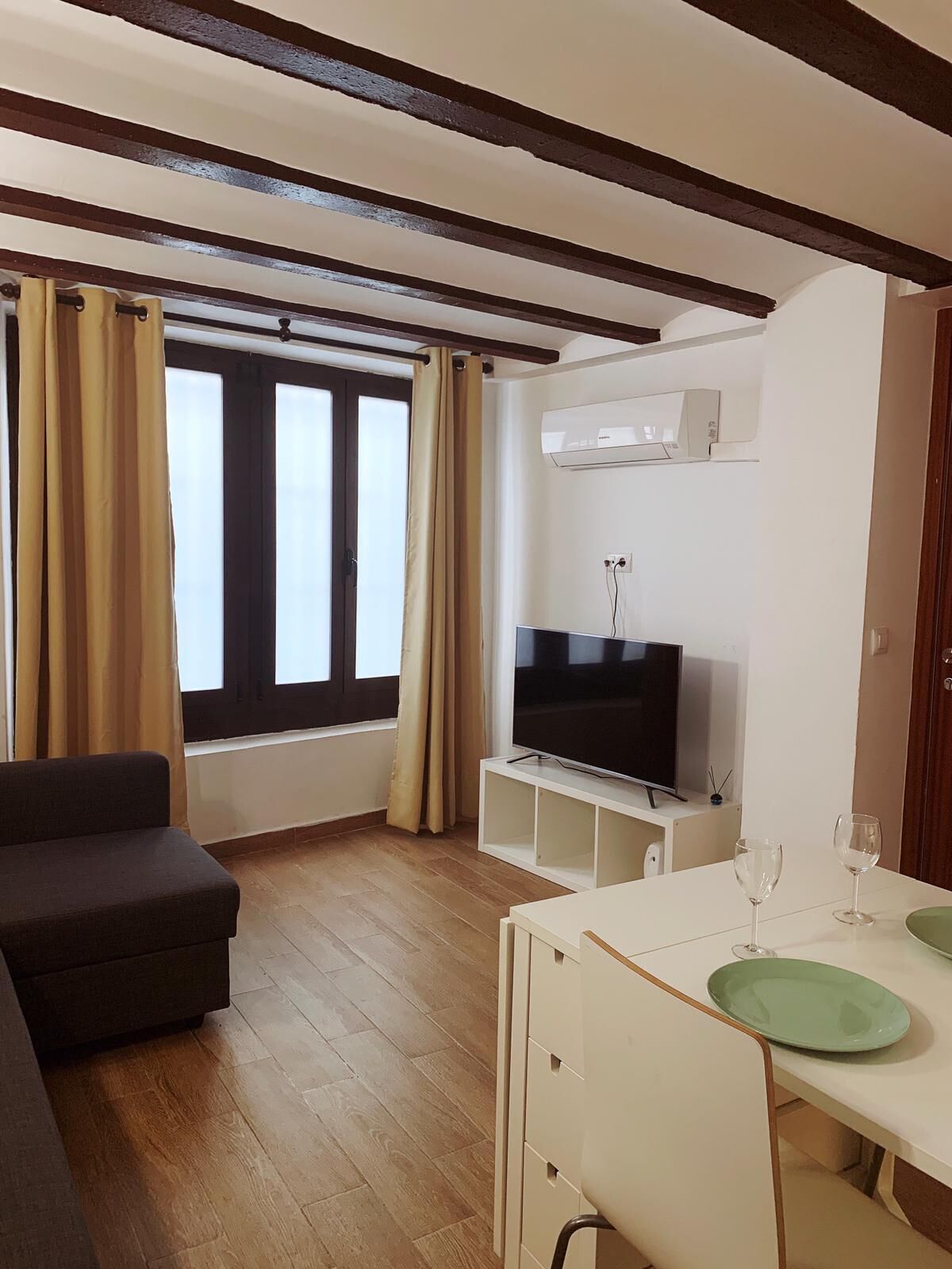 A living room with a sofa, a TV on a stand, a dining table with glasses, and a window with curtains.