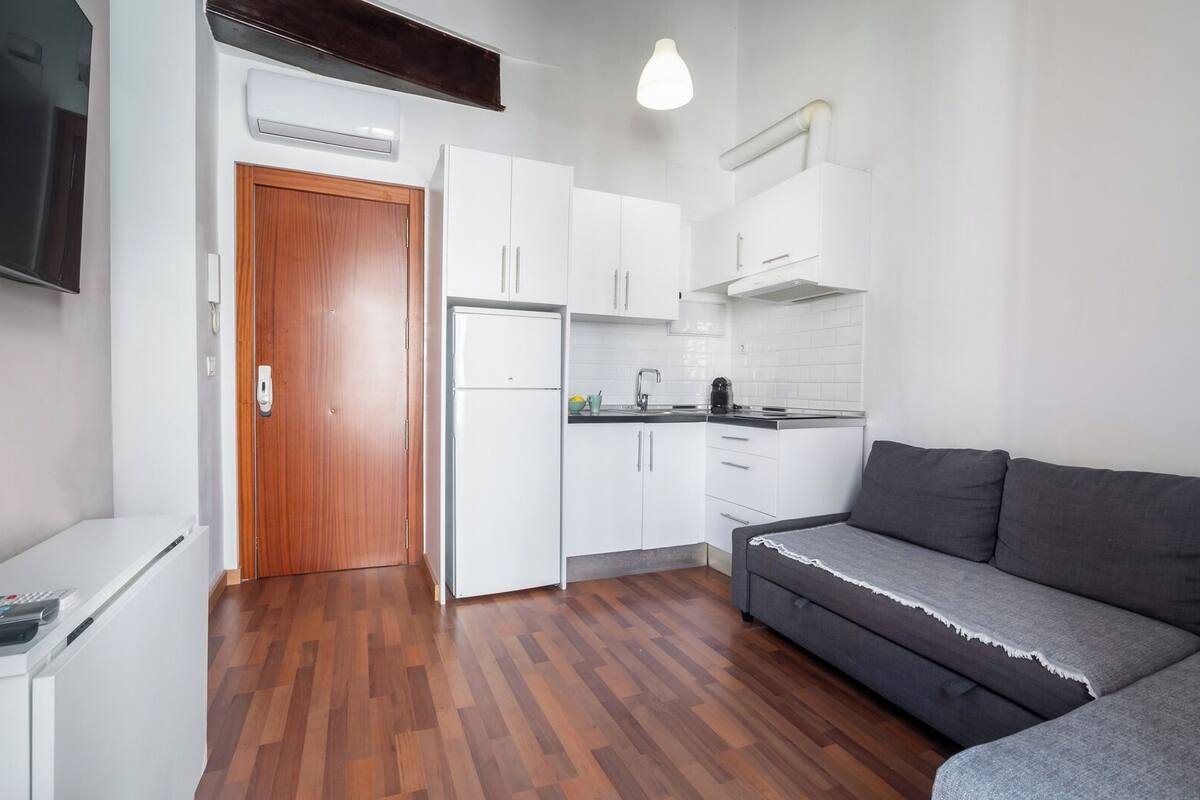 Eine kleine K üche mit weißen Schränken, einem eingebauten Backofen und einem grauen Sofa.