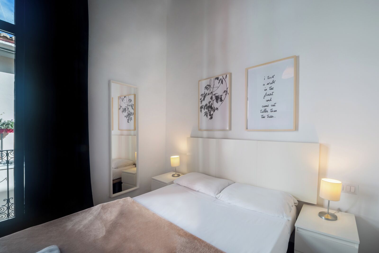 Hotellihuoneessa on sänky, kaksi yöpöytää, joissa on lamput, suuri peili ja kaksi kehystettyä kuvaa seinällä.