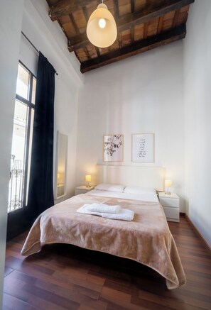 Un dormitorio con techo de madera, una cama grande con colcha color beige, dos mesitas de noche con lámparas, y dos cuadros enmarcados en la pared.