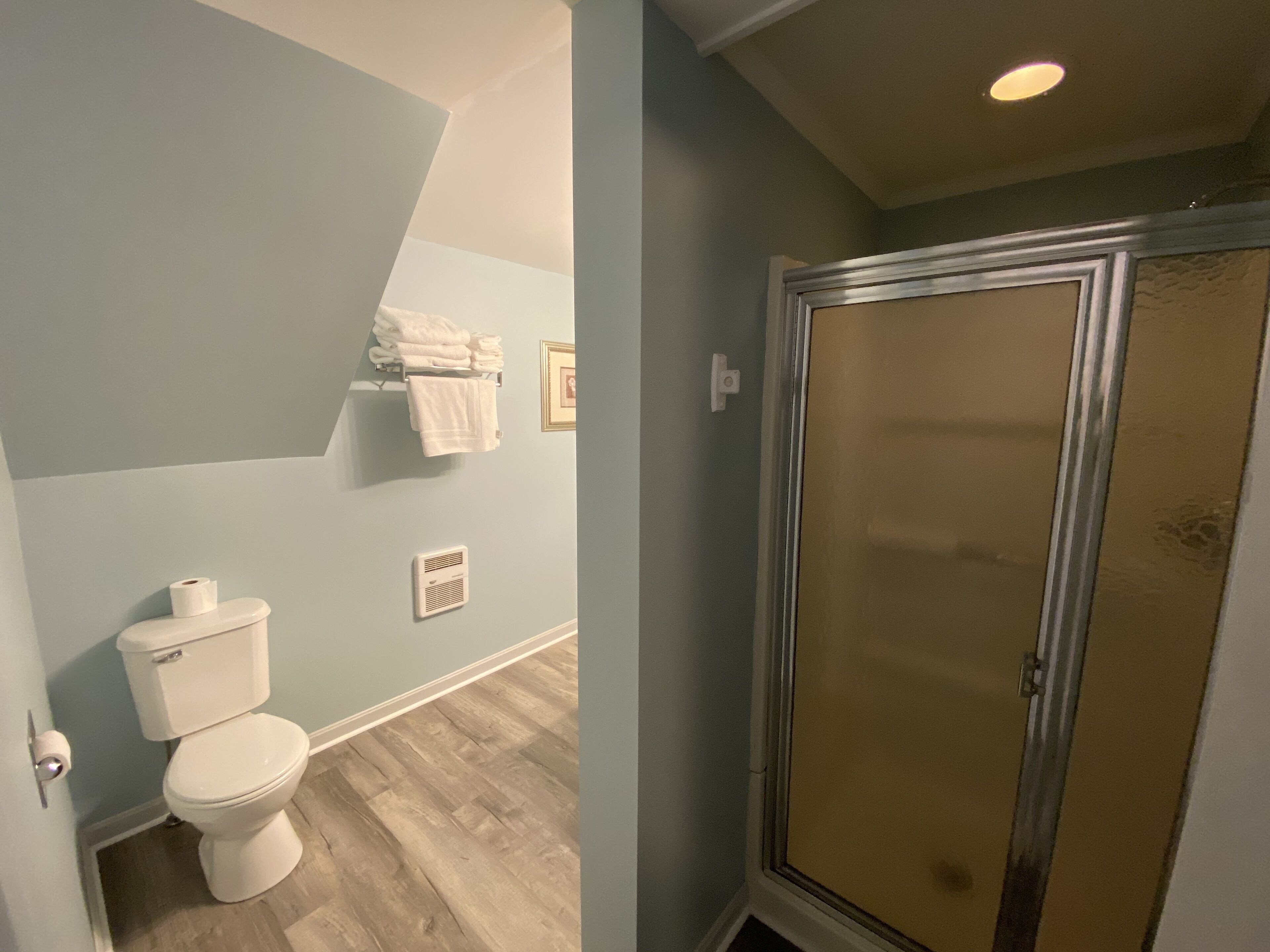 A bathroom with a toilet, a towel rack with folded towels, a vent, and a shower with a glass door.