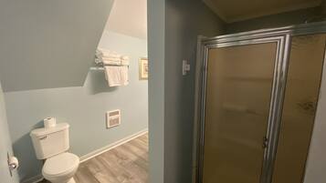 A bathroom with a toilet, a towel rack with folded towels, a vent, and a shower with a glass door.