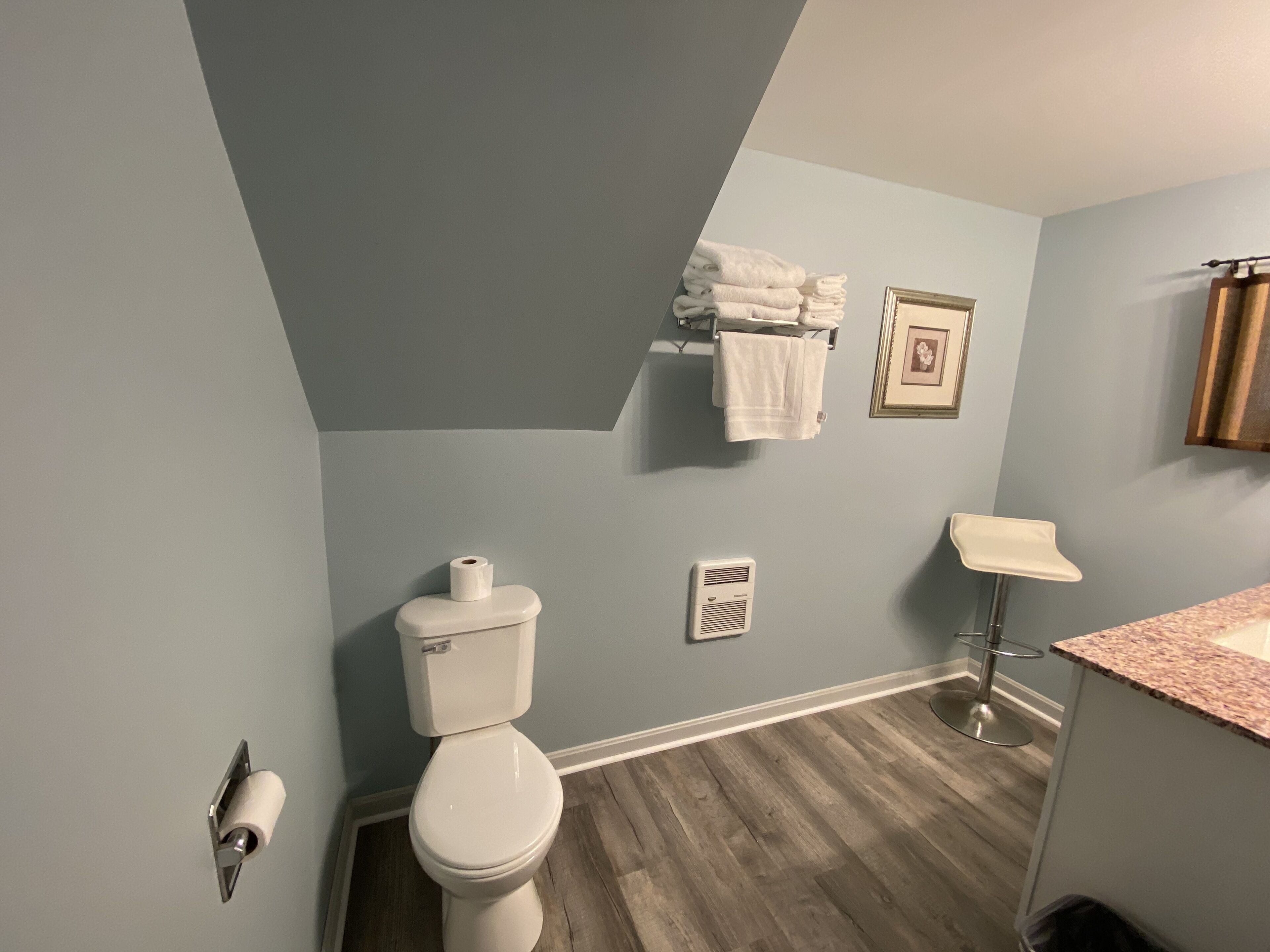 A bathroom with a toilet, a towel rack with towels, a picture frame, and a shower chair.