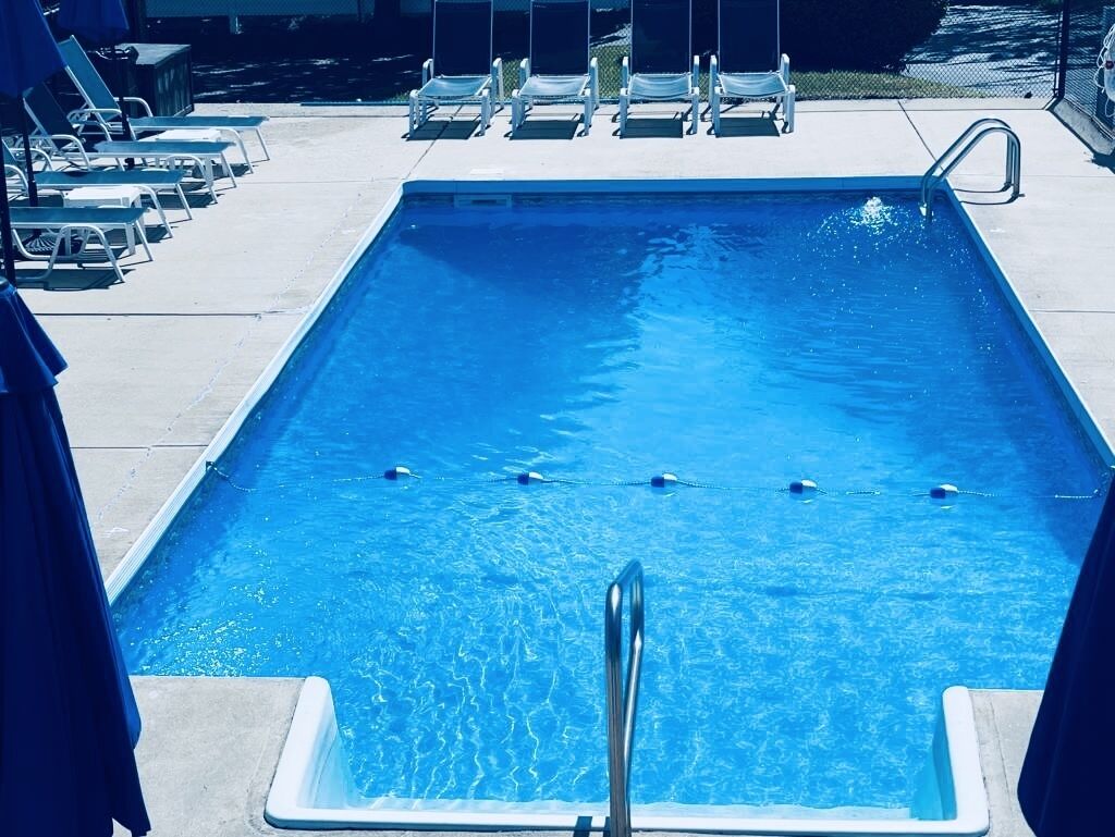 Piscina retangular com escada metálica na parte rasa, cercada por espreguiçadeiras e guarda-sóis.