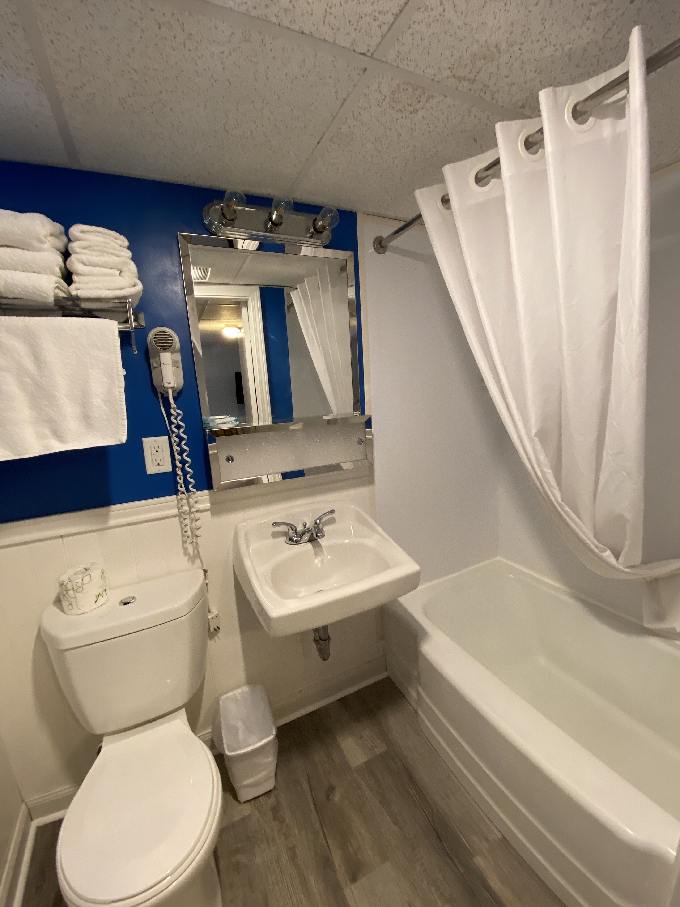 Baño con inodoro, lavamanos y bañera. Hay un perchero con toallas, un secador de pelo y un cubo de basura.