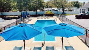 En swimmingpool med blå parasoller och solstolar, omgiven av ett staket och träd.
