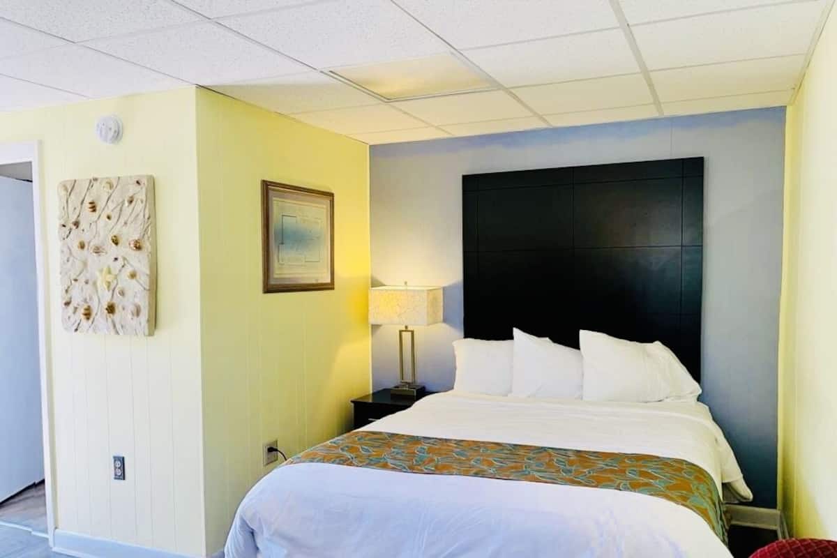 A hotel room with a large bed, a nightstand with a lamp, a wall-mounted artwork, and a framed picture.
