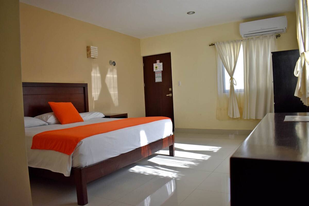 A hotel room with a bed, a nightstand, an air conditioning unit, and a door with a sign.