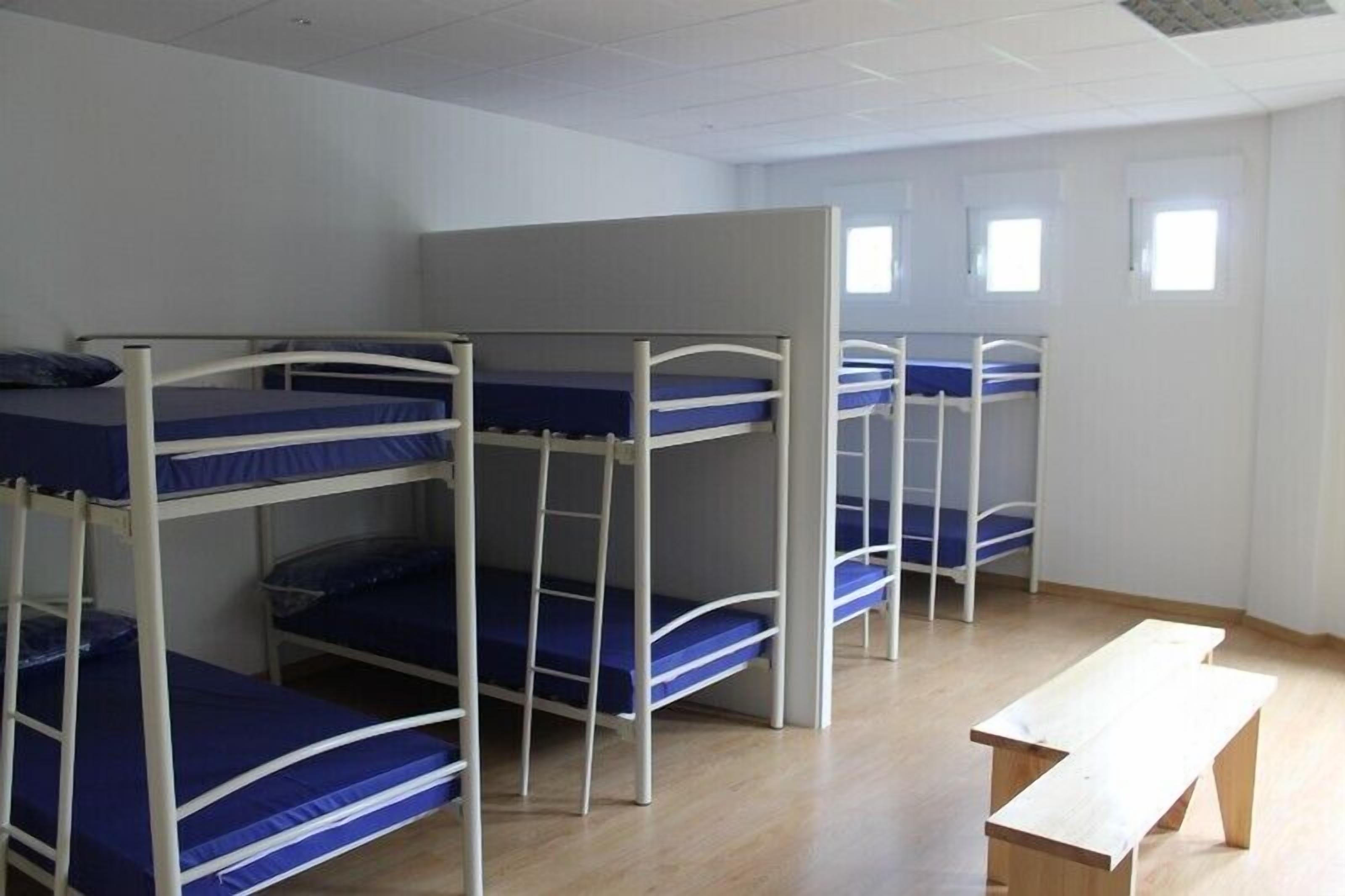 A dormitory room with bunk beds, a wooden bench, and a window.