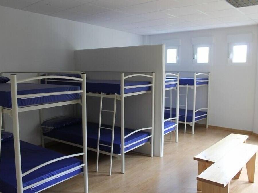 A dormitory room with bunk beds, a wooden bench, and a window.