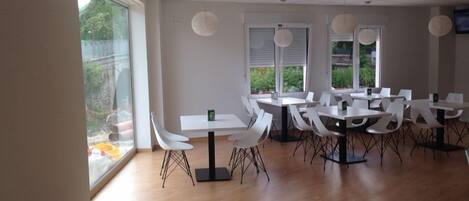 A spacious room with multiple white tables and chairs, large windows with blinds, and a flat-screen TV mounted on the wall.