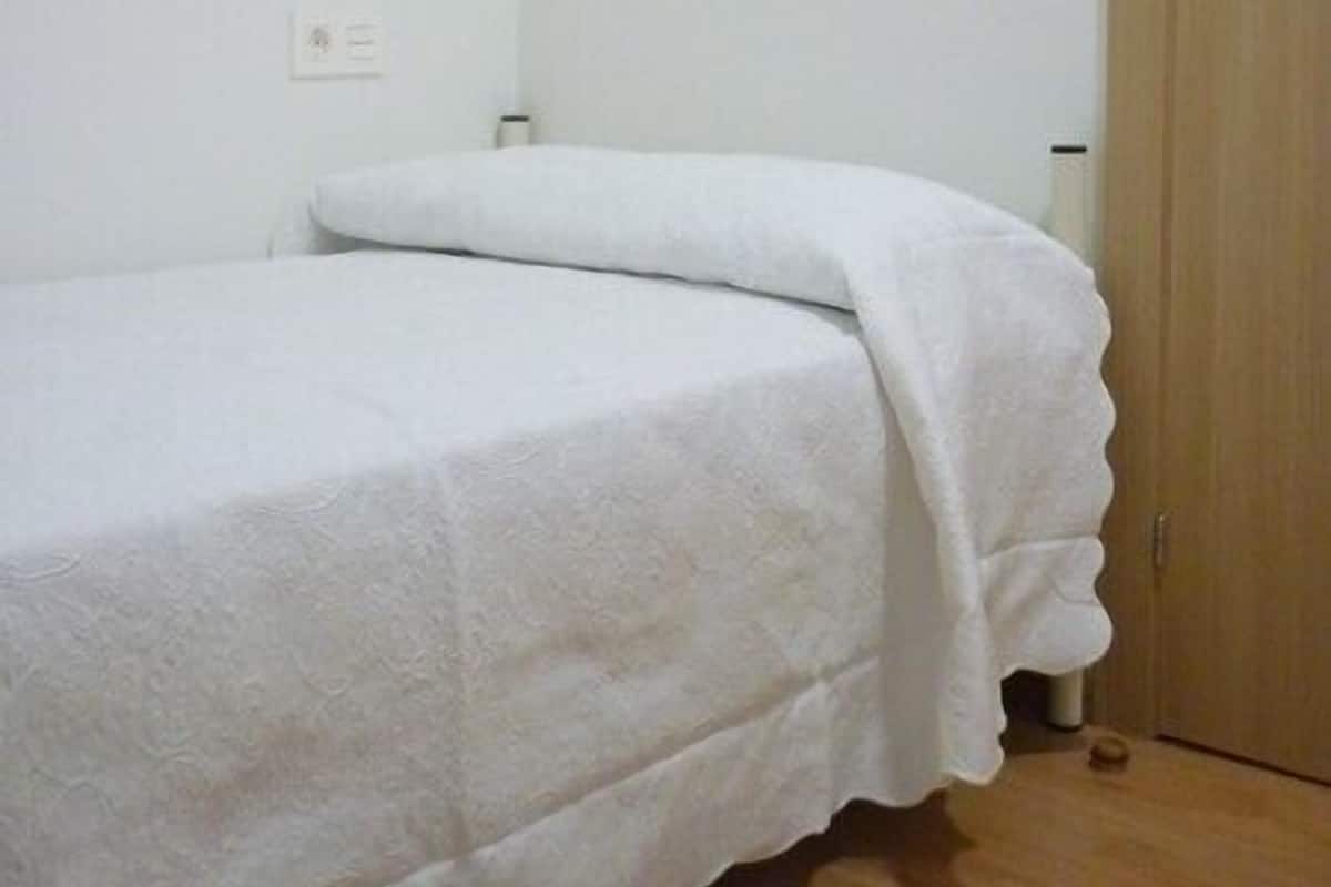 A single bed with a white bedspread in a room with a wooden wardrobe and a white wall.