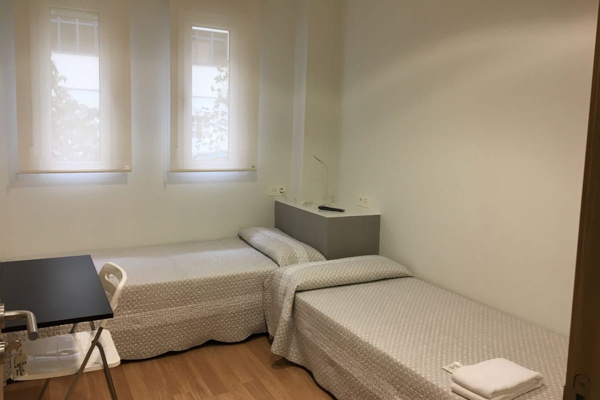 A small, clean room with two single beds, a desk, and a chair. There are two windows providing natural light.
