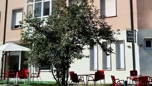 A building with a stone wall, red chairs, and a tree in front. - Pensión Albergue Puente Ribeira - Hostel (Sarria)