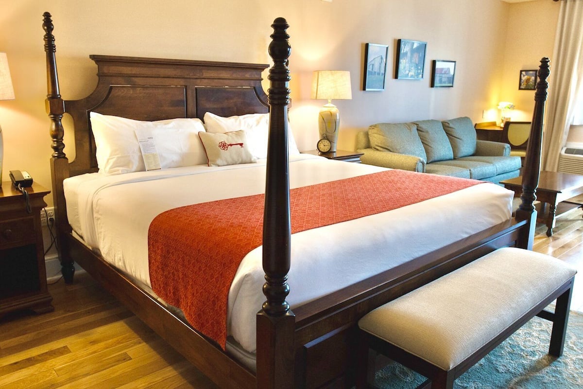 Una cama con dosel con un plaid rojo, una mesita de noche de madera con una lámpara, y un banco al pie de la cama.