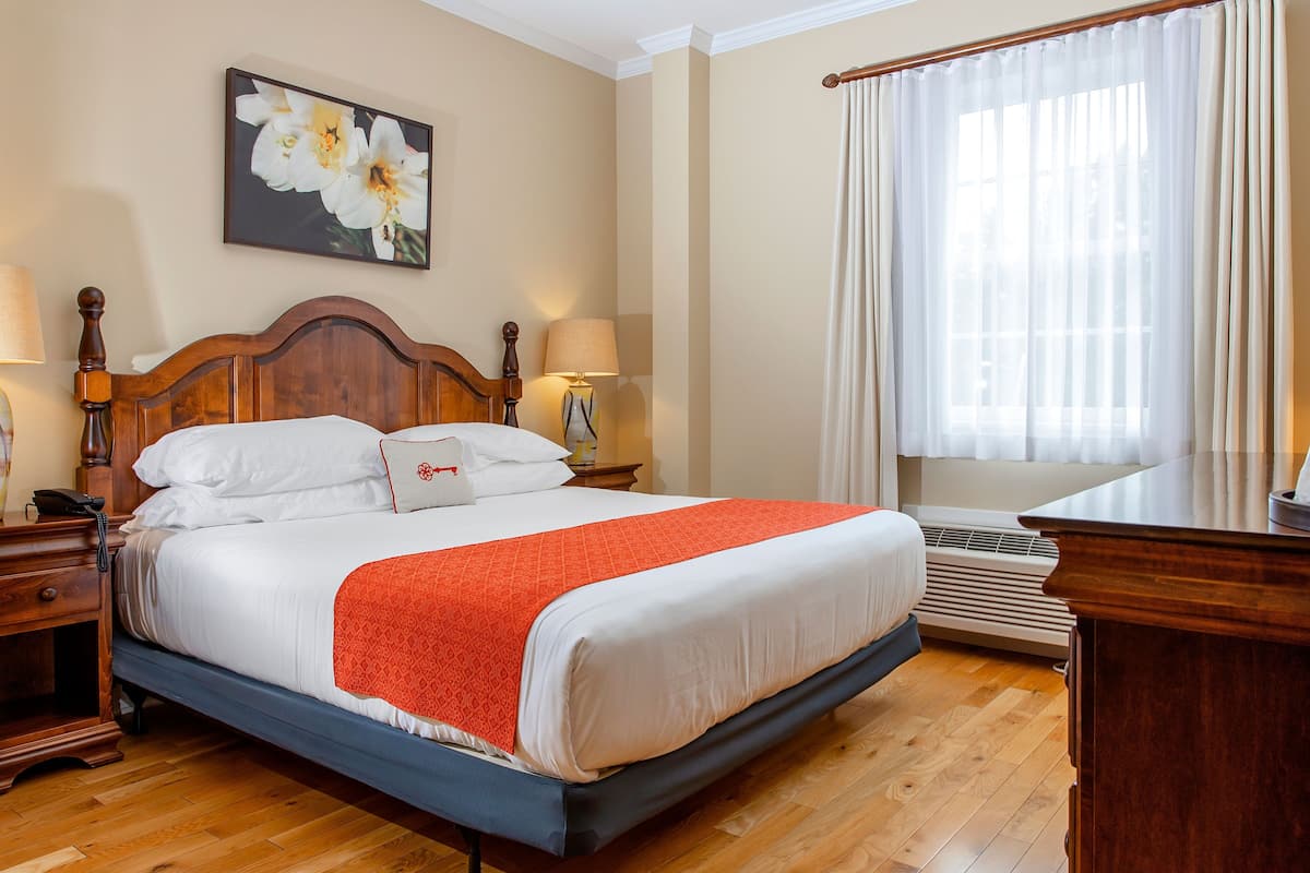 Habitación de hotel con una cama grande, cabecera de madera, dos mesitas de noche con lámparas, un aparador y un ventanal con cortinas.