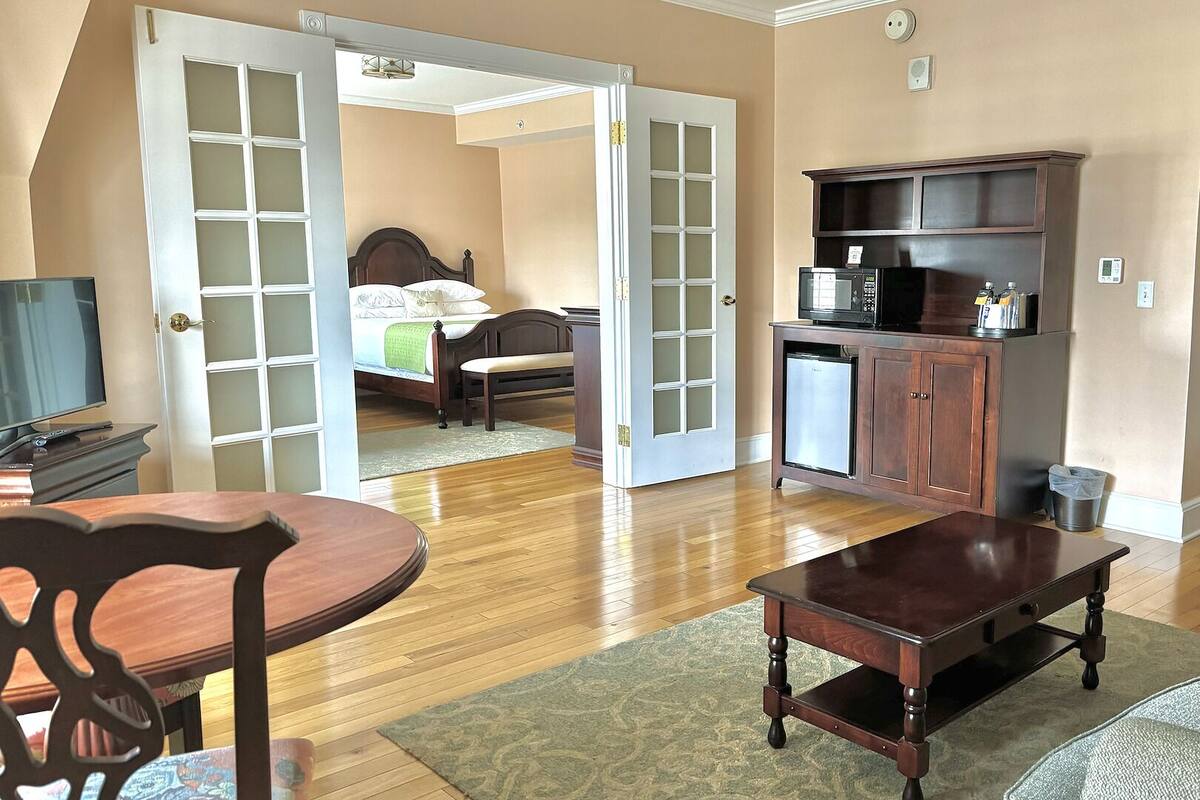 Una habitación amplia con suelo de madera, una cama con cabecero, una pequeña kitchenette, una mesa de comedor con sillas y un televisor.