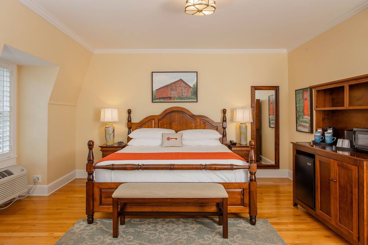Un dormitorio con cama de madera, lámparas de noche, una mesita de noche pequeña, un banco, televisión empotrada y una zona de cocina con microondas y armarios.