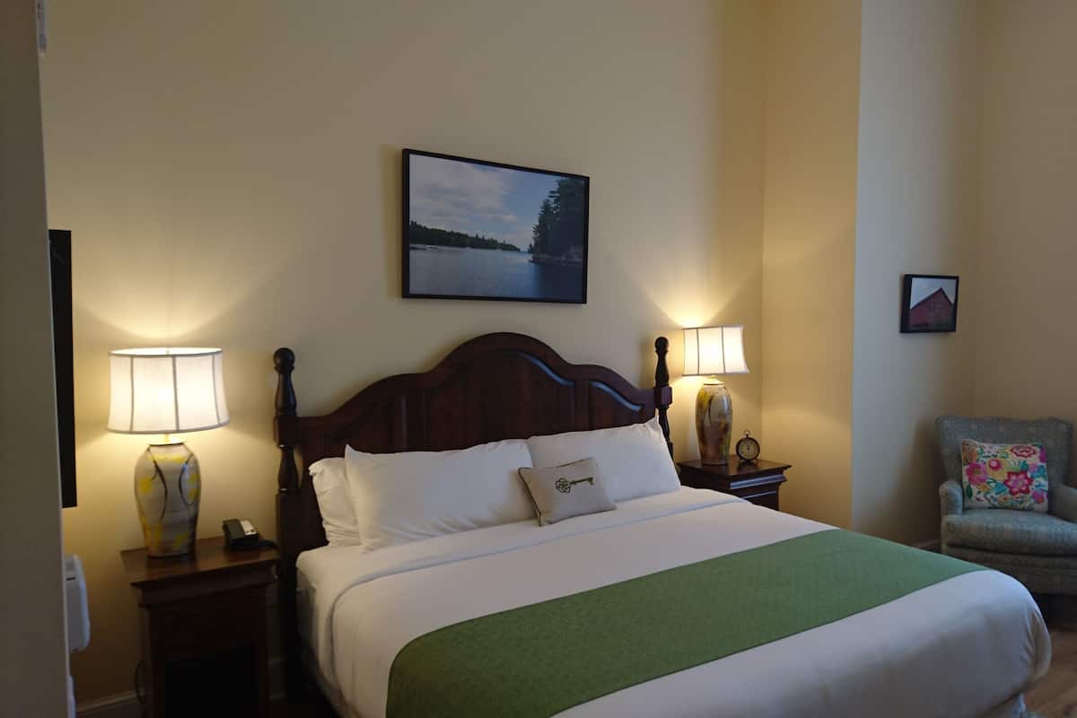 Una habitación de hotel con una cama, dos lámparas de noche, una silla, un cuadro en la pared y una ventana con cortinas.