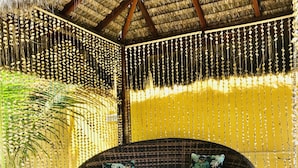 Un canapé rond en osier agrémenté de coussins à motifs floraux, un toit de chaume et des rideaux de perles suspendues, le tout dans un espace extérieur baigné de soleil.