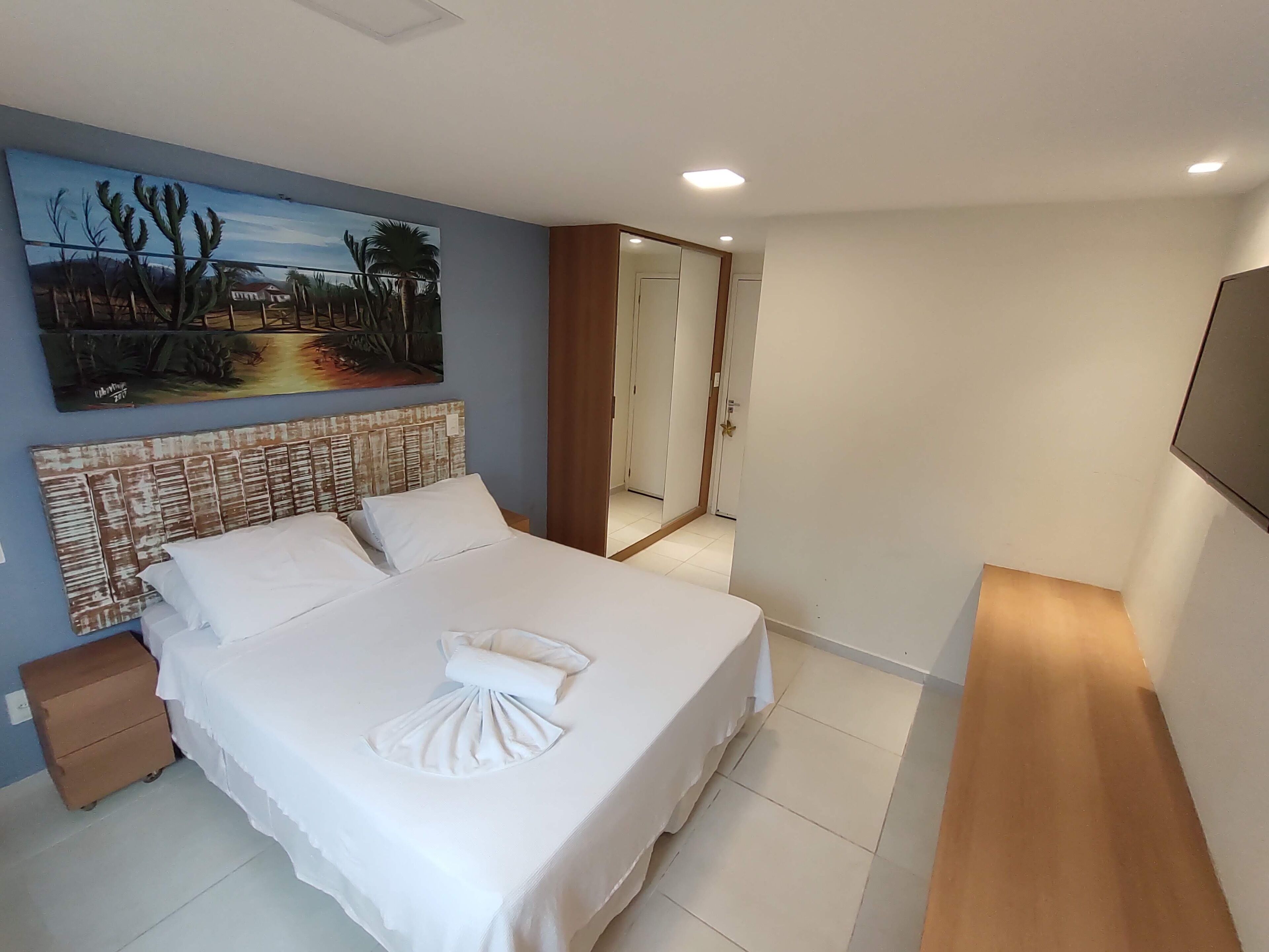 A neatly made bed with white linens and a towel, a painting of a landscape on the wall, and a wooden floor.