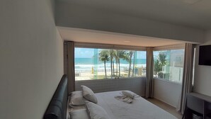 Une chambre avec une grande fenêtre donnant sur une plage, un lit avec du linge de maison blanc et un téléviseur à écran plat fixé au mur.