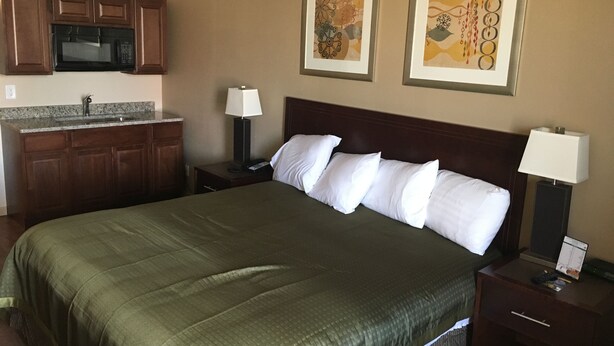 A hotel room with a bed, a nightstand, a lamp, a desk with a lamp, a microwave, and a sink.