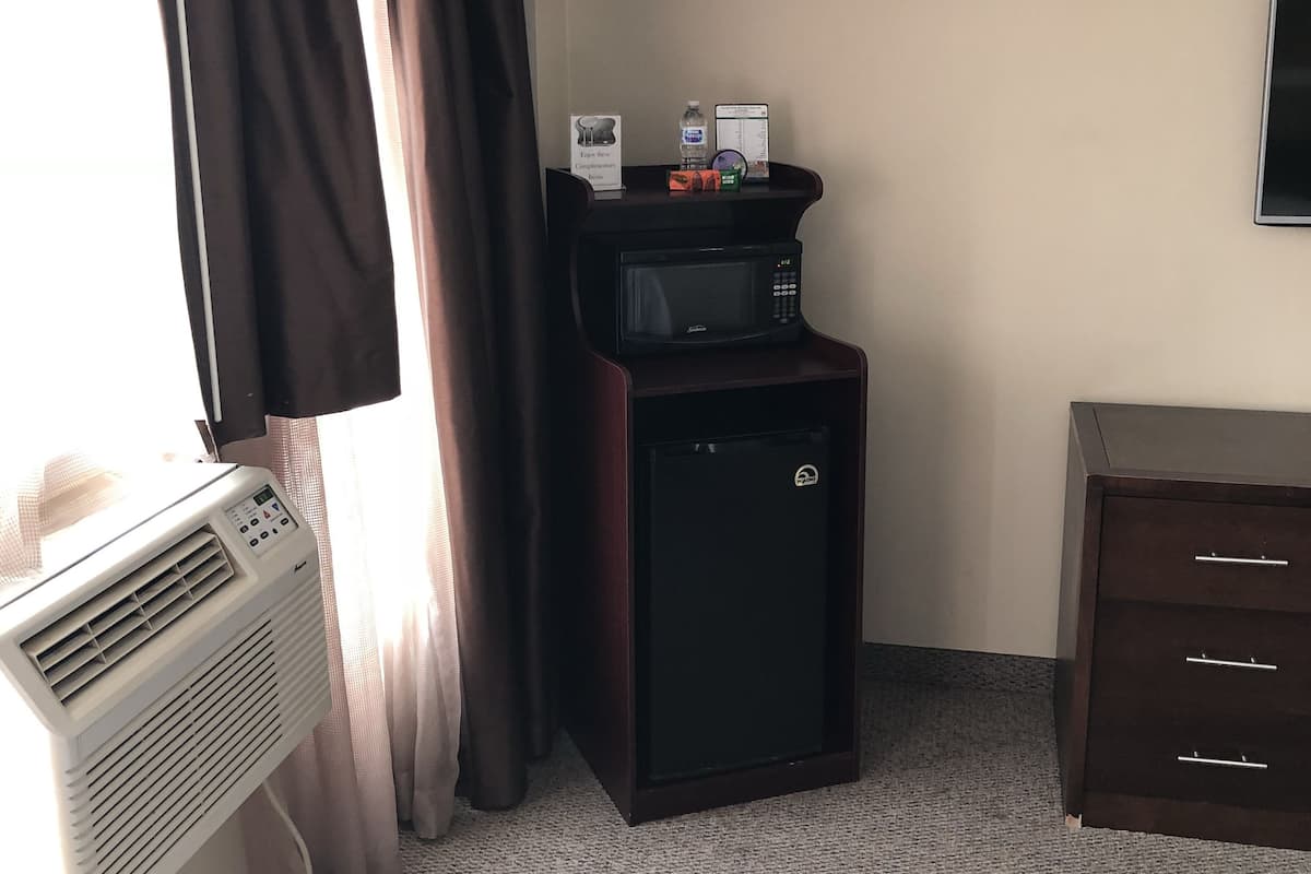 A hotel room with a window, curtains, a wall-mounted TV, a small black refrigerator, a desk with a water bottle, a coffee maker, and a microwave.