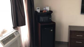 A hotel room with a window, curtains, a wall-mounted TV, a small black refrigerator, a desk with a water bottle, a coffee maker, and a microwave.