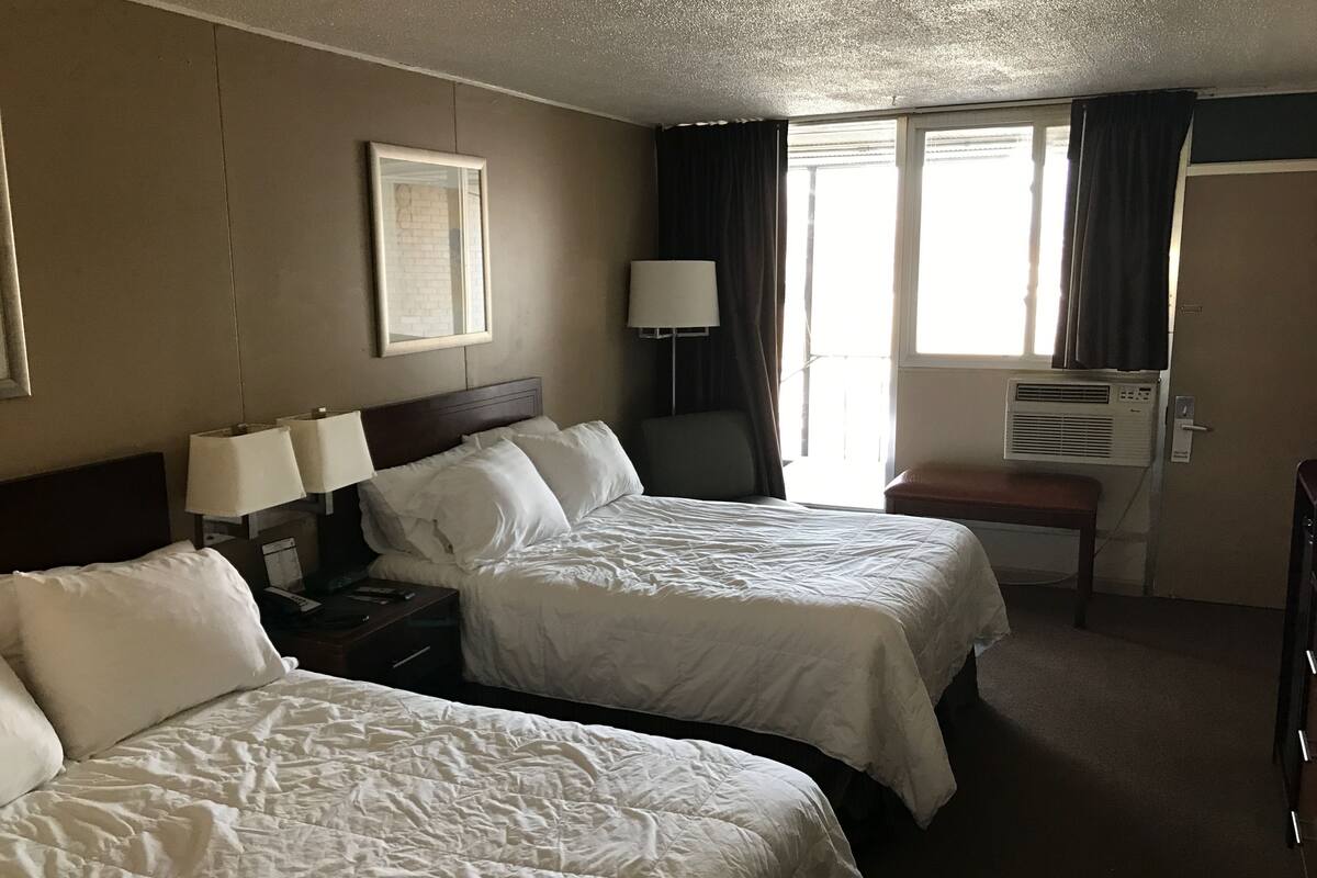 A hotel room with two beds, a desk, a chair, a window with curtains, and an air conditioning unit.