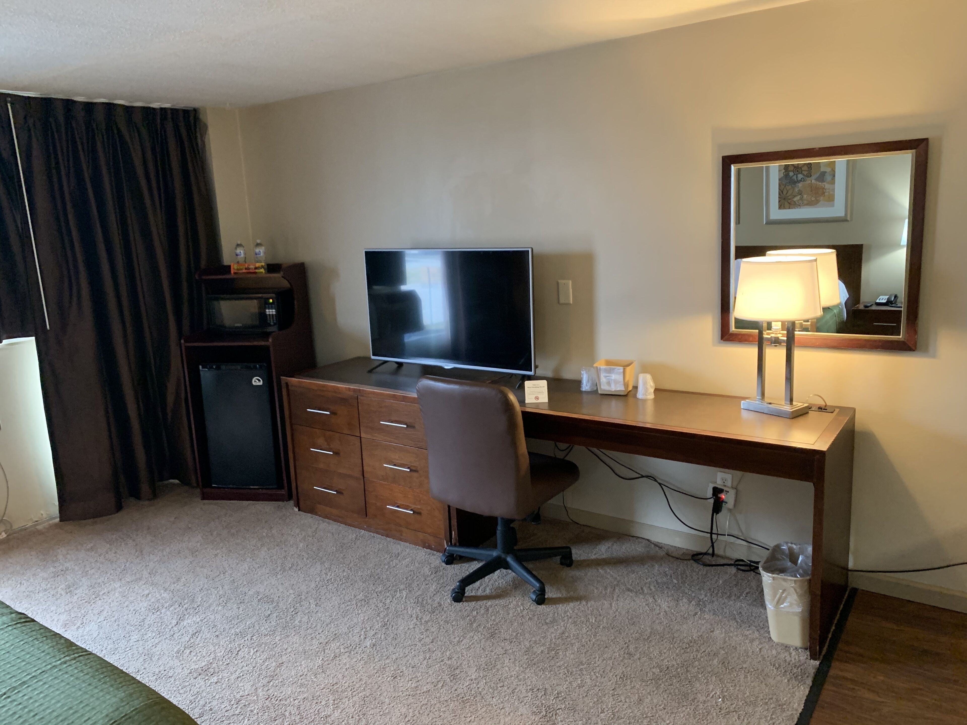 A hotel room with a wooden desk, a television, a chair, a lamp, a trash bin, and a mirror on the wall.