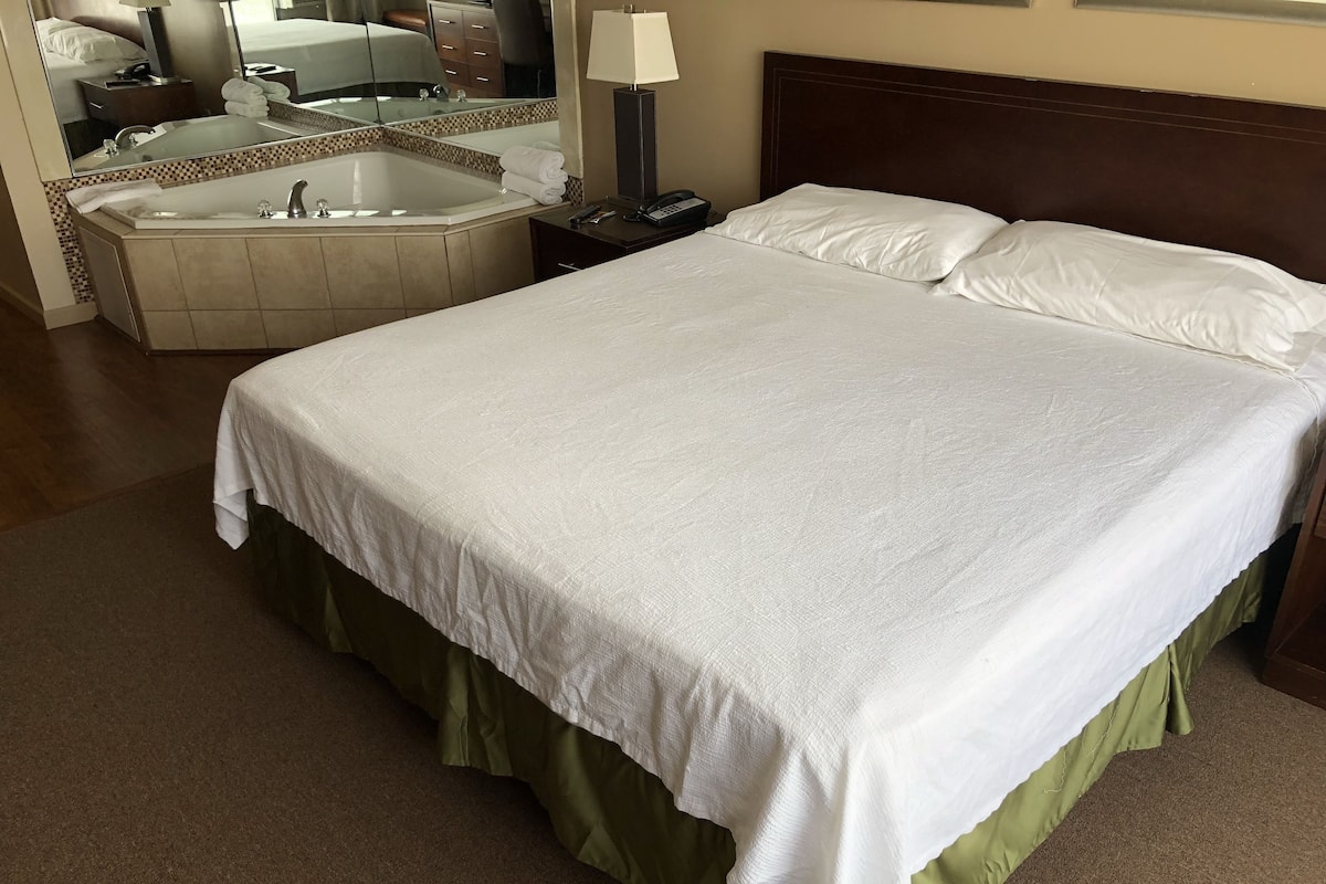 A hotel room with a large bed, a separate bathroom with a bathtub, a mirror, and a bedside table with a lamp.