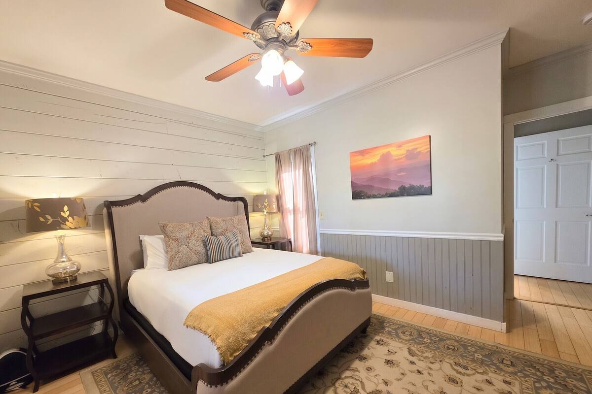 Quarto com cama, mesa de cabeceira, luminária, ventilador de teto, quadro e porta.