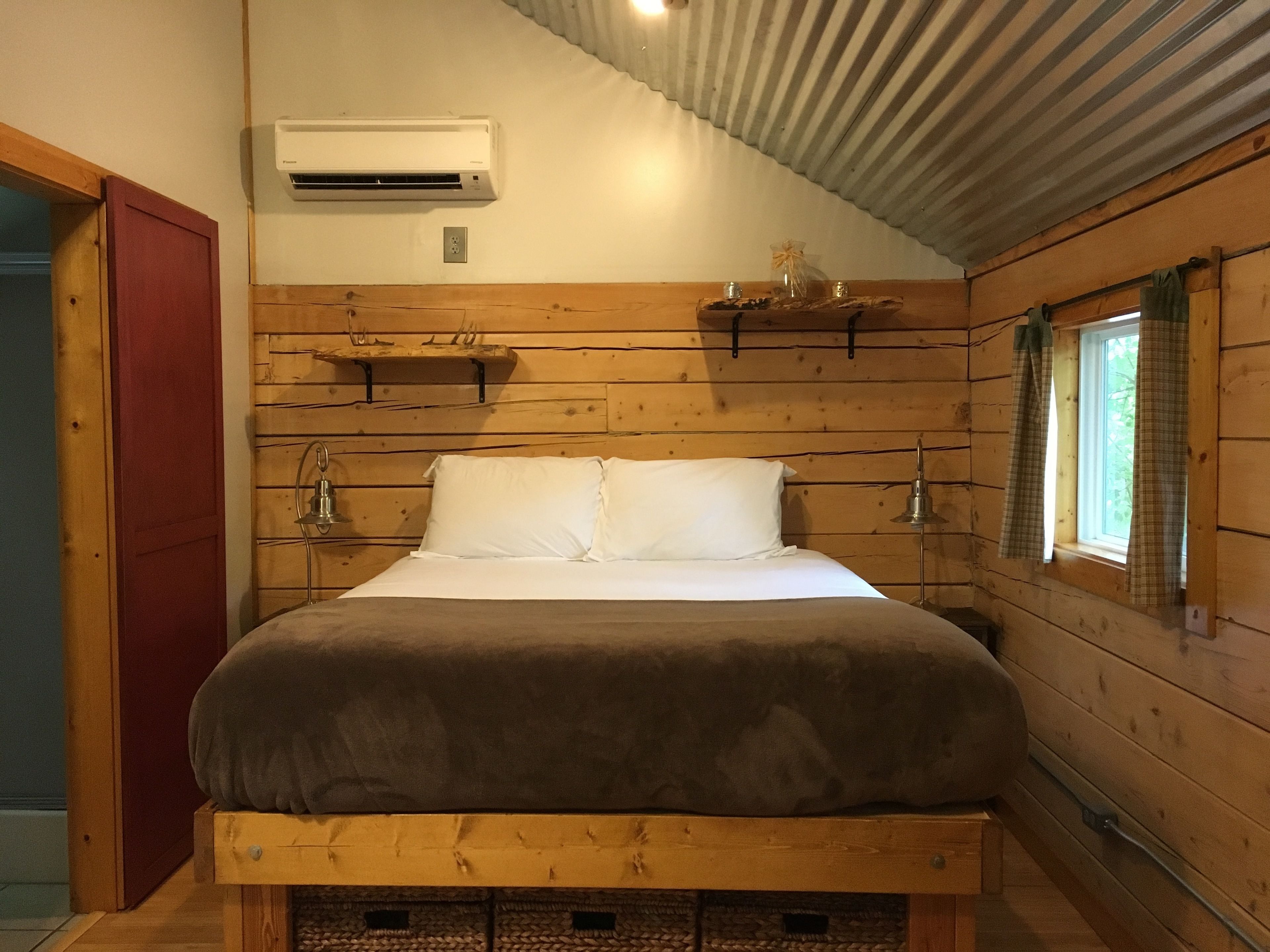 A wooden cabin bedroom with a bed, a shelf with decorative items, a window with curtains, and an air conditioning unit.