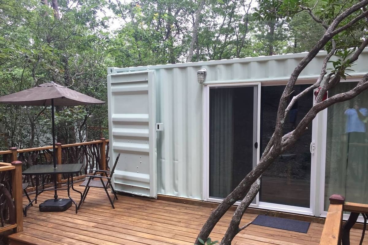 Uma construção compacta e térrea, com porta de correr de vidro, um pequeno deck com mesa e cadeiras e um guarda-sol de patío.