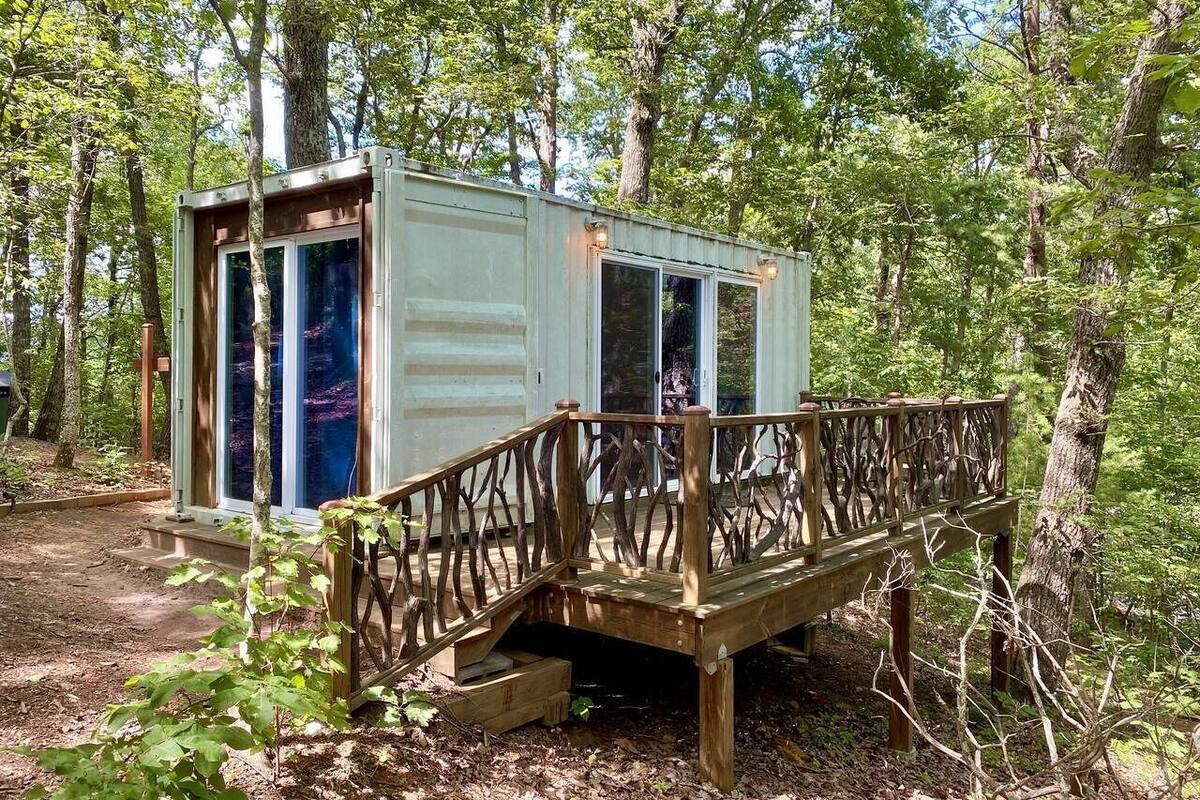 Uma cabana pequena com deck de madeira e portas de vidro, rodeada por árvores.