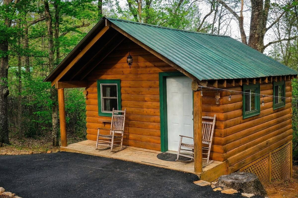 Uma pequena cabana de toras com telhado verde, duas cadeiras de madeira na parte externa e uma porta.