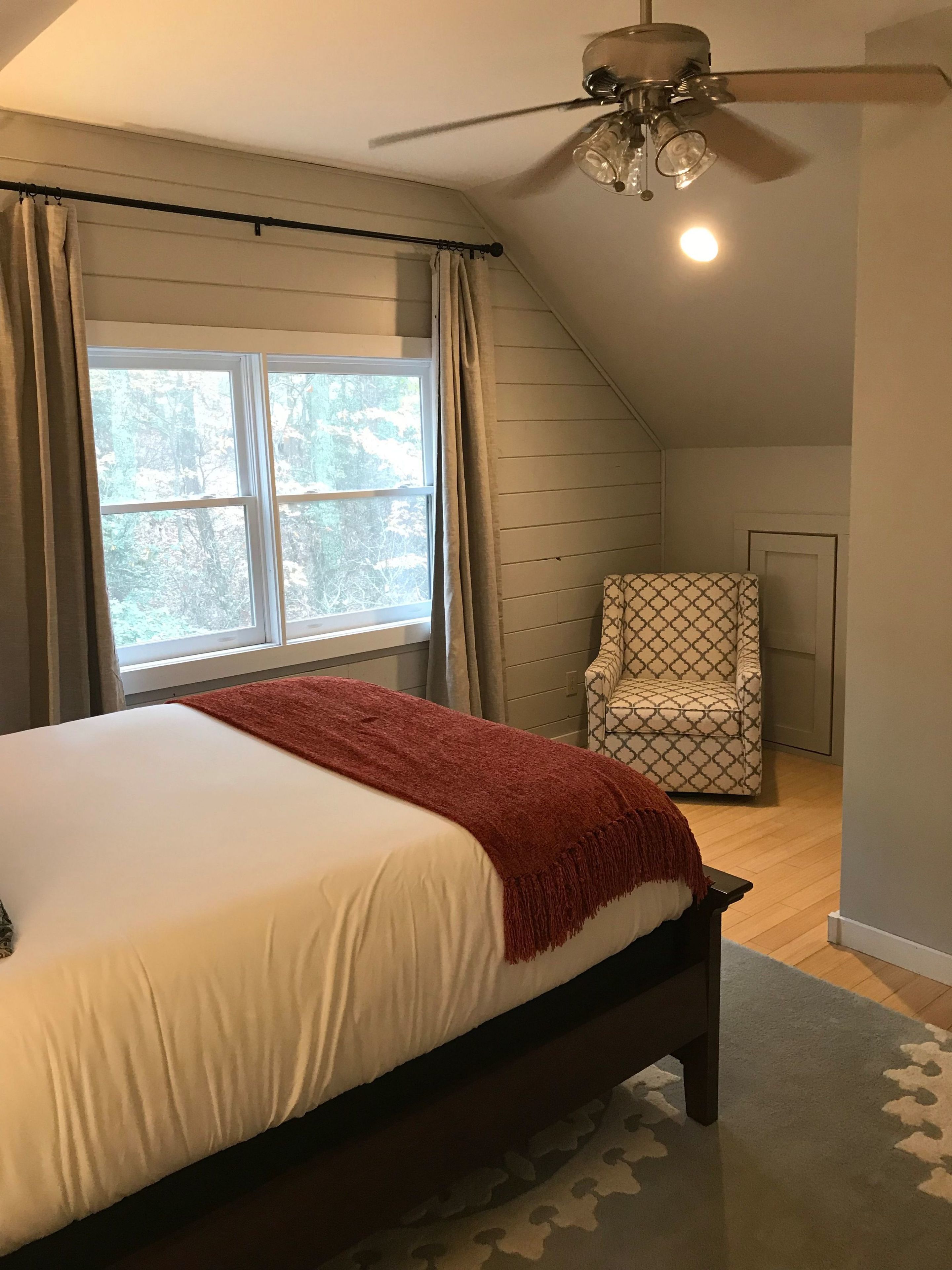 A bedroom with a bed, a ceiling fan, a window, and a patterned chair.