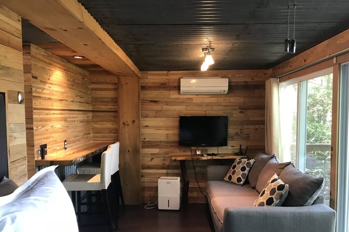 Um quarto acolhedor em estilo de cabana, com decoração em madeira, uma TV de tela plana na parede, um sofá com almofadas estampadas e uma grande janela que proporciona luz natural.